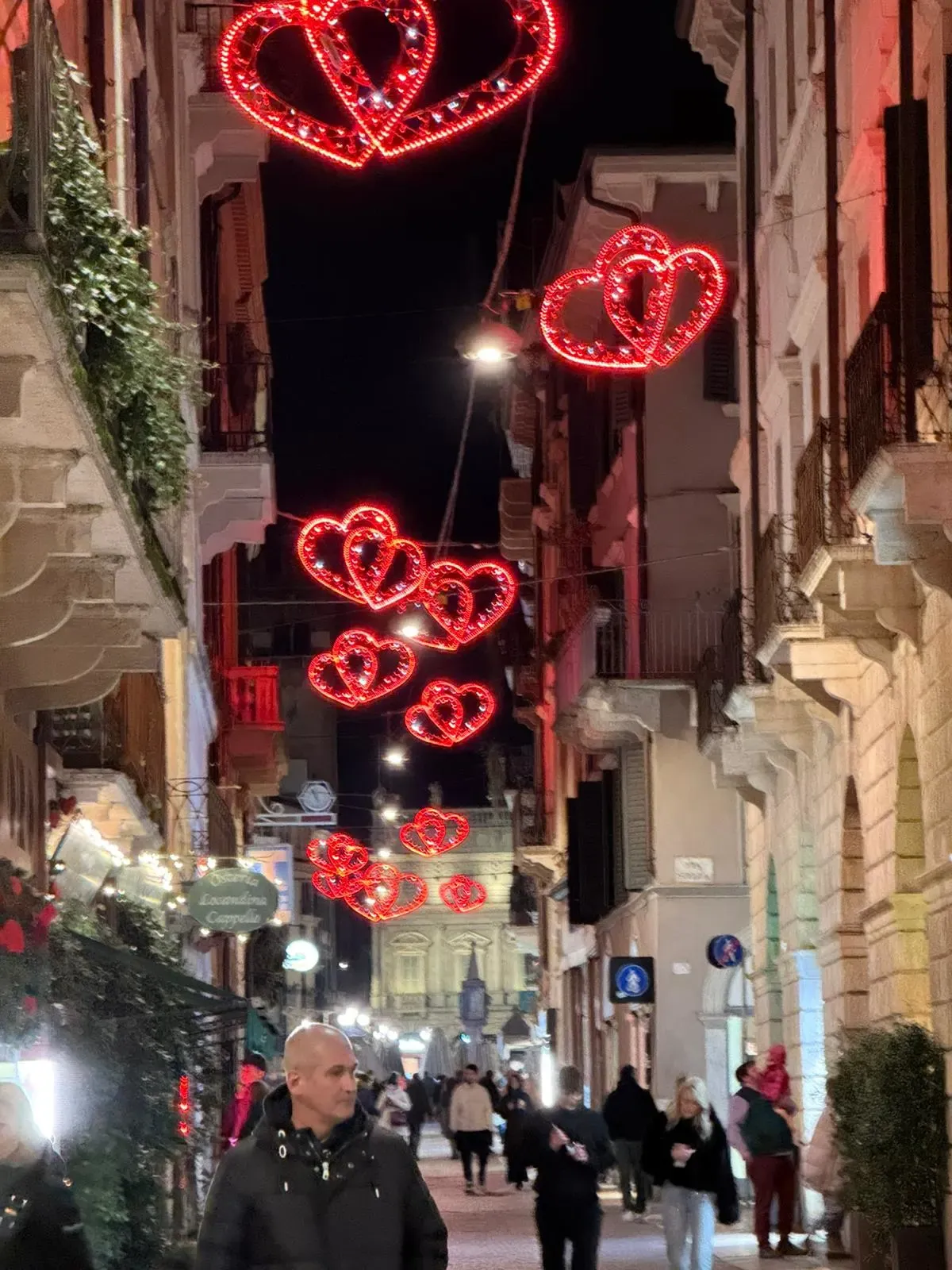 A lively street scene in Verona, Italy, decorated with bright red heart-shaped lights hanging above for Valentine's Day. People are walking along the cobblestone street, surrounded by historic buildings and storefronts, creating a festive and romantic atmosphere at night.