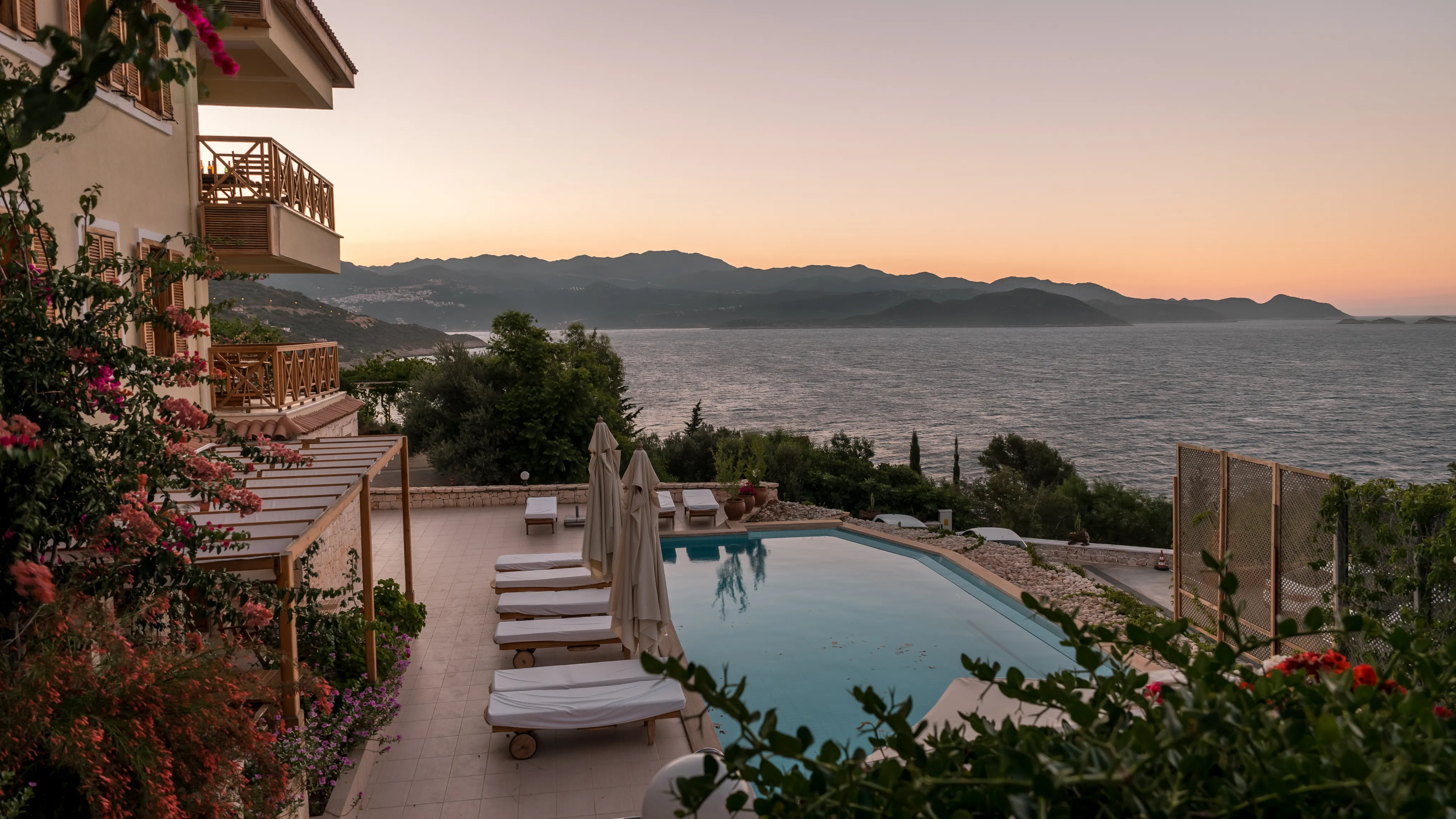 Scenic view of a serene poolside terrace overlooking the sea at sunset, surrounded by lush greenery and mountains in the background.
