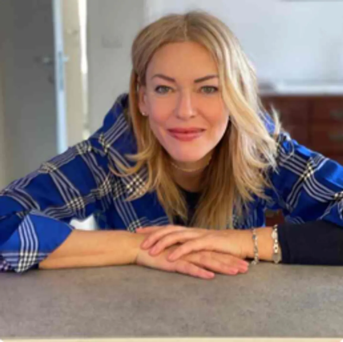 A woman (Antonella Fraccaro, host) with long blonde hair wearing a blue plaid shirt is leaning forward on a countertop, smiling warmly at the camera. The background shows a partially open door and a kitchen area.