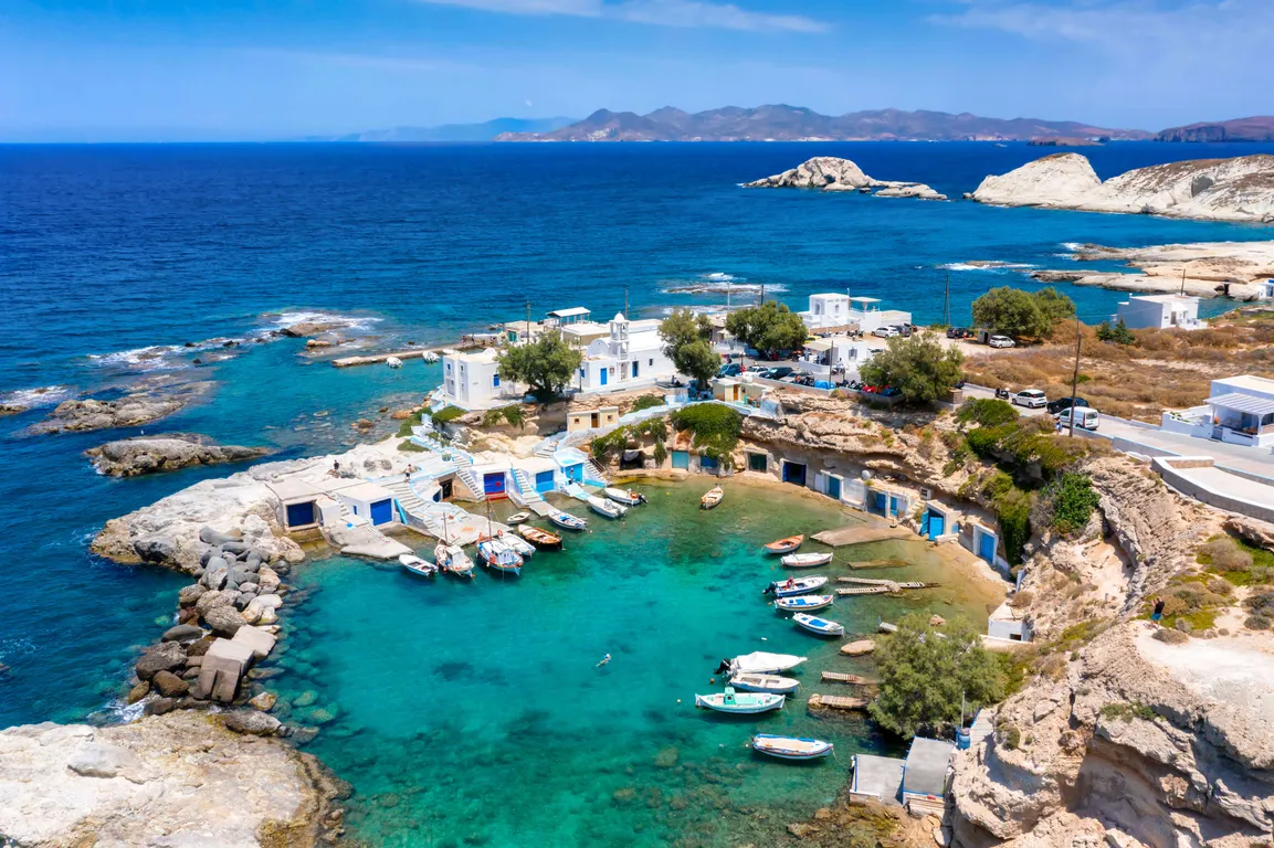 A picturesque coastal village with whitewashed buildings and blue doors sits along a small, turquoise bay surrounded by rocky cliffs. Small fishing boats are docked in the calm water, and a few people can be seen swimming and walking along the shore. The deep blue Aegean Sea stretches out in the background, with distant islands and rugged rock formations dotting the horizon.